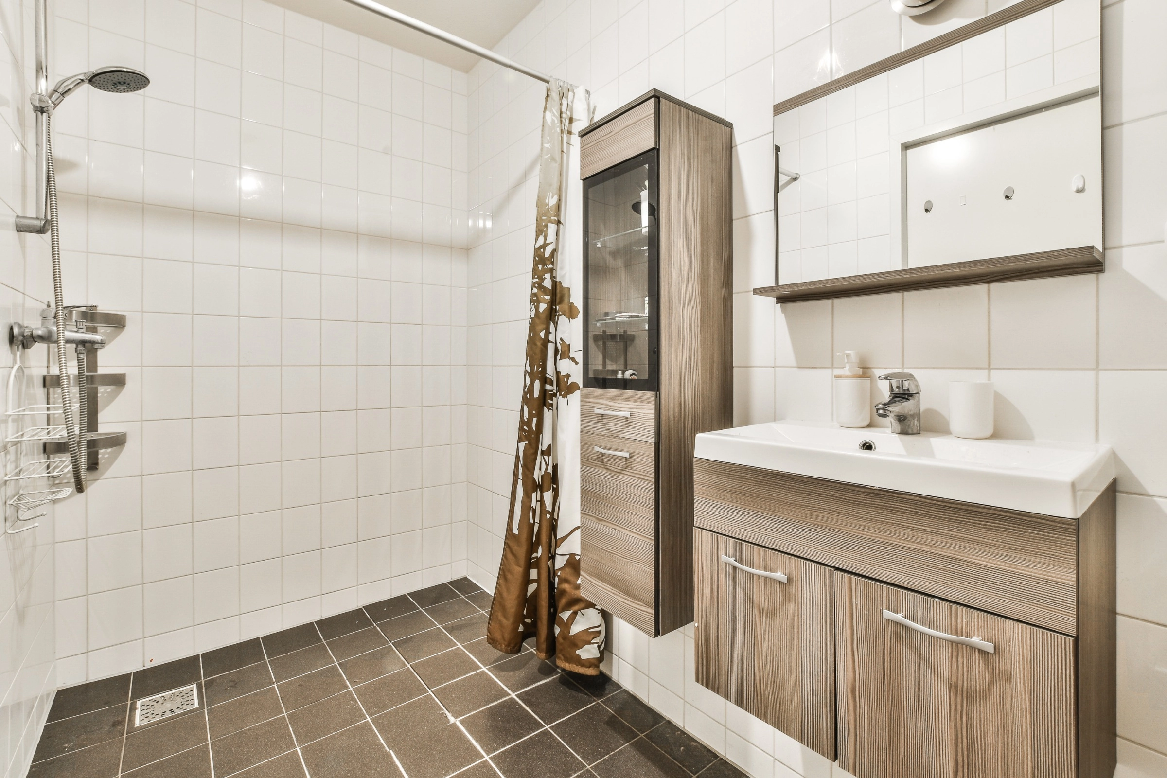 A small, basic bathroom with white tiled walls, a shower with a brown floral curtain, a wooden vanity cabinet with a single white sink, a tall wooden storage cabinet, a mirror above the sink, and dark brown floor tiles.